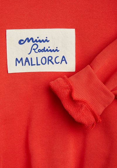 Világos piros ruha fehér folttal, amelyen a „mini Rodini MALLORCA” felirat látható, és fodros ujjmandzsetta jobb oldalon.