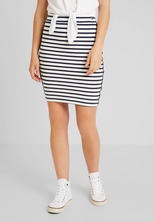 Kvinde iført nederdel med navyblå og hvide vandrette striber, hvidt top med knude foran og hvide hæsthøjtop-sneakers på ensfarvet baggrund.