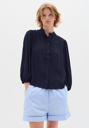 Blouse bleu marine avec un col haut, fermeture à boutons et manches bouffantes, associée à un short bleu clair avec un ourlet décoratif.
