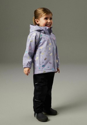 Usměvavé dítě v světle fialové bundě s kapucí a barevnými potisky jednorožců, v černých kalhotách a černých botách, stojící na šedém pozadí.