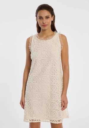 Frau trägt ein ärmelloses, knielanges cremefarbenes Kleid mit strukturierten Blumenmustern und transparenten, gerafften Verzierungen an Schultern und Ausschnitt.