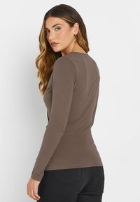 Langärmliges, runder Hals hochwertiges Oberteil aus gedämpftem braunen Stoff. Figurbetontes Design mit glatter Textur und minimalen Nähten. Kombiniert mit dunklen Jeans.