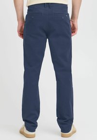 Temno modri chinos hlače s ravnim krojem, dvema zadnjima žepoma s gumbi in gladko bombažno teksturo. Nosi se z svetlejšimi čevlji.