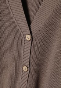 Bruine geribbelde cardigan met een V-hals en een enkele houten knoopsluiting. De textuur is gebreid met verticale lijnen en een zachte afwerking.