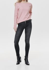 Różowy sweter z dzianiny z okrągłym dekoltem, zestawiony z czarnymi obcisłymi dżinsami i czarnymi botkami na obcasie. Dżinsy mają dopasowany krój.