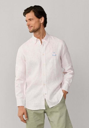 Homme portant une chemise rose clair à manches longues avec un petit logo bleu sur la poitrine, associée à un pantalon vert clair, regardant sur le côté.