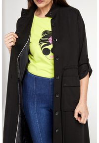 Cappotto lungo nero con chiusura a bottoni, caratterizzato da una tasca frontale e una fodera interna. Indossato sopra una camicia verde brillante con un motivo grafico.