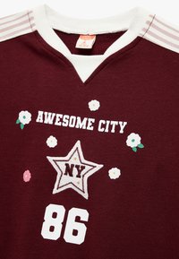 Bordeauxfarbener Sweater mit weißem V-Ausschnitt. Mit den Aufschriften "AWESOME CITY", einem Stern mit "NY" und floralen Applikationen. Strukturierter Stoff; gerippte Bündchendetails.