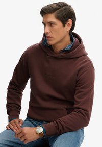 Felpa bordeaux con tasca a canguro e logo ricamato. Indossata sopra una camicia a quadri blu, abbinata a jeans in denim chiaro e un orologio dorato.