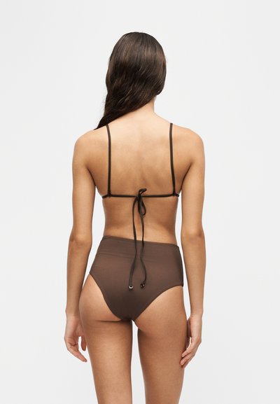 Femme aux longs cheveux foncés portant un bas de bikini marron taille haute et un haut de bikini assorti noué dans le dos, debout devant un fond uni.