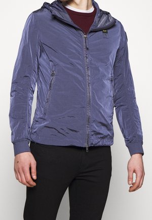 Mężczyzna ubrany w niebieską lekką, zapinaną na zamek kurtkę z kapturem na bordową koszulę i czarne spodnie, stojący na jednolitym tle.