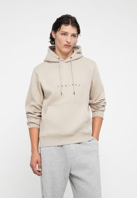 Ung mand i beige hættetrøje med forlommer og snore, iført lysegrå sweatpants, stående med den ene hånd i lommen mod hvid baggrund.