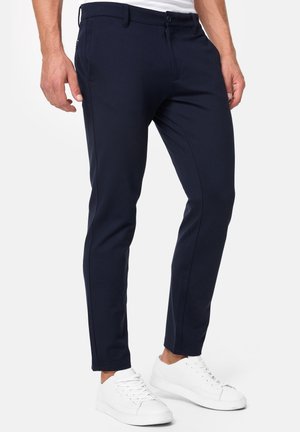 Mann trägt schlanke marineblaue Hose und weiße Sneakers, steht vor einem schlichten hellen Hintergrund.