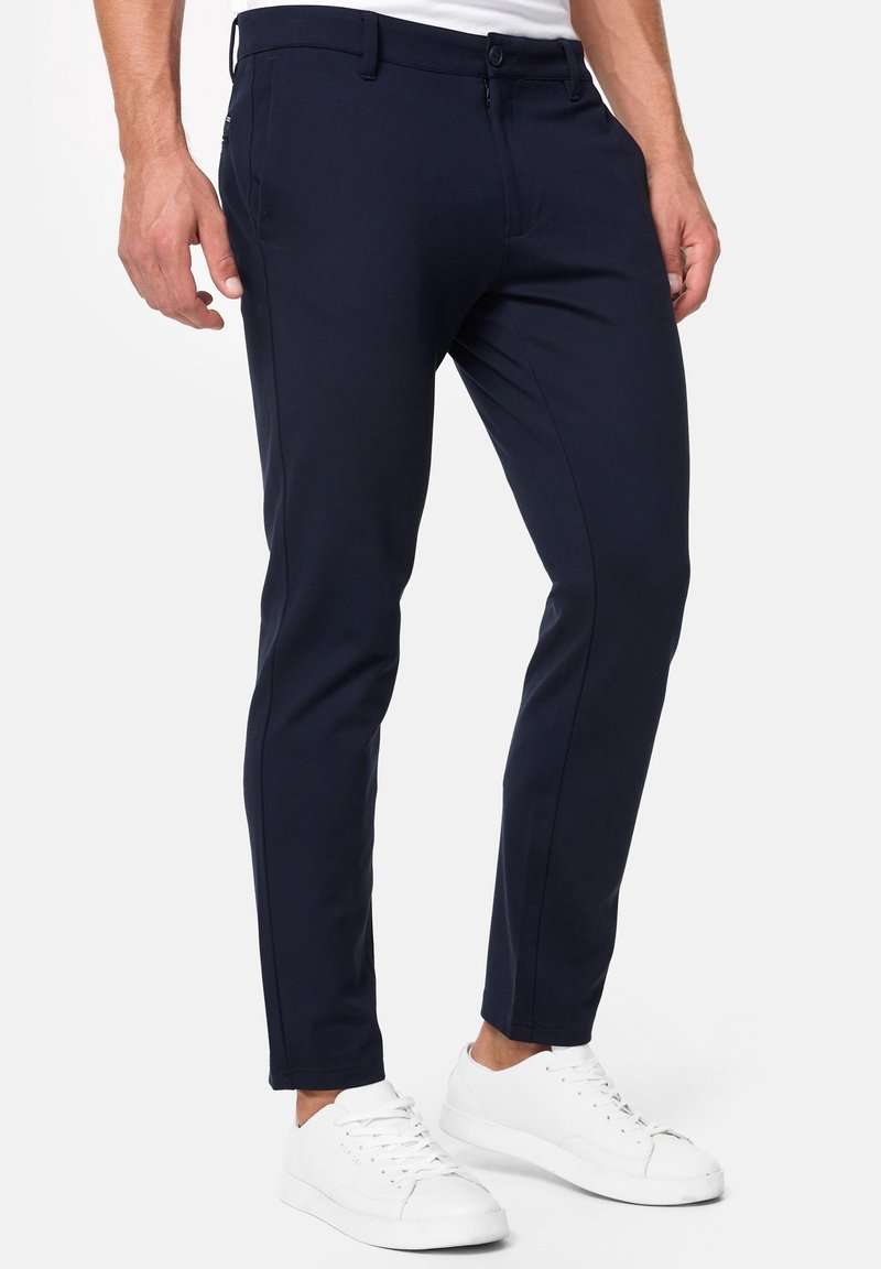 Homme portant un pantalon bleu marine slim et des baskets blanches, debout devant un fond clair uni.