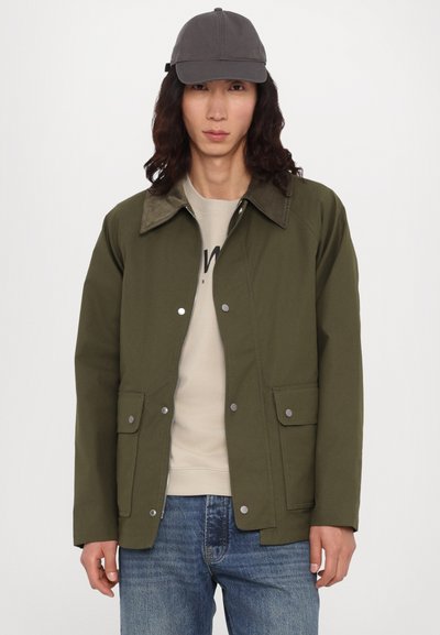 Veste verte au design uni, avec un col marron contrasté et des boutons-pression, associée à un haut beige et un jean bleu.