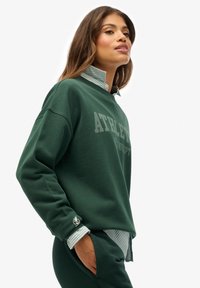 Sudadera verde oscuro con el texto "ATHLETIC", que presenta un cuello a rayas. El material parece suave. Incluye un detalle del logotipo en el puño.