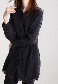 Camicia a maniche lunghe, blu scuro, con bottoni, orlo sfilacciato, in morbido cotone, con vestibilità rilassata e spacchi laterali per facilitare i movimenti.