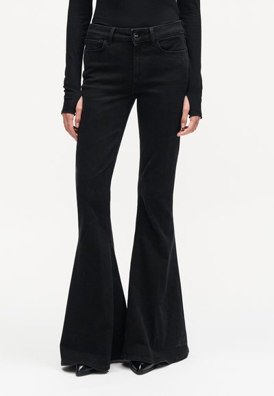 Jean noir taille haute évasé porté avec des chaussures noires à bouts pointus et un haut noir à manches longues, visible de la taille jusqu'aux pieds.