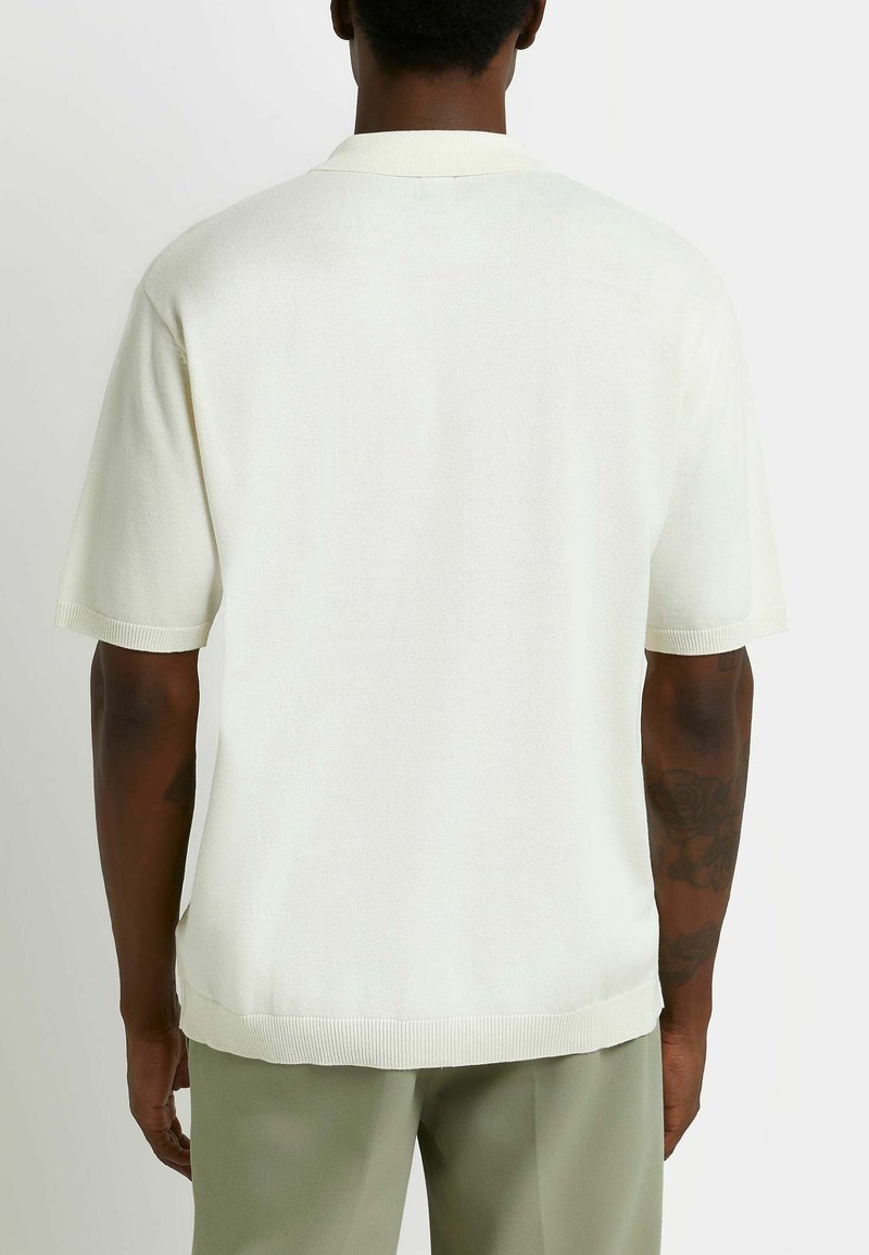 Stickad pikétröja i off-white med mjuk textur, korta ärmar, ribbad krage och fåll. Sedd bakifrån, visar en avslappnad passform.