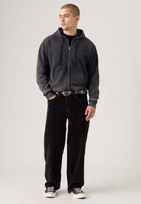 Hooded zip-up sweatshirt in washed black, paired with black corduroy pants and black sneakers with white soles. Belt features circular accents.