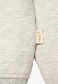 Zbliżenie beżowej dzianiny z małą kremową metką z napisem "BILKVIS" w jasnobrązowym kolorze.