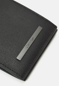 Zwarte leren portemonnee met een textuurafwerking, zilverkleurig metalen accent met "Calvin Klein" gegraveerd en zichtbare stikdetails.