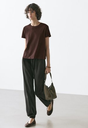 Frau mit lockigen Haaren, die ein braunes Kurzarmoberteil, weite schwarze Hosen, braune Ballerinas trägt und eine grüne Handtasche hält.