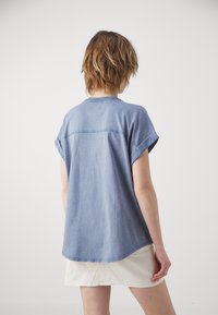 Cămașă din denim albastru, cu mânecă scurtă și decolteu rotund, având manșete răsucite, cu o cusătură orizontală pe spate. Fabricată dintr-un amestec moale de bumbac.