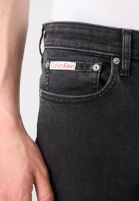 Vaqueros de mezclilla negra con un bolsillo frontal que lleva la etiqueta de Calvin Klein, herrajes plateados y un acabado texturizado.