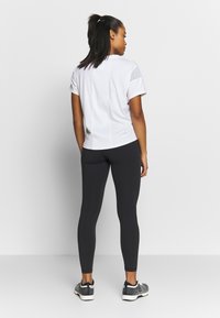 T-shirt atlético branco com mangas curtas, combinado com leggings pretas. A modelo está a usar ténis cinzentos com um design texturizado.