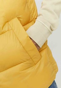 Chaqueta acolchada amarilla con una textura suave y un bolsillo lateral, llevada sobre una manga acanalada de color claro. Diseño compacto y ligero.