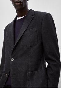 Uomo che indossa un blazer grigio scuro con tasche a toppa sopra un maglione scuro e una maglietta bianca, su uno sfondo chiaro e uniforme.