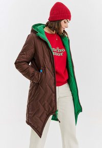 Prešívaný hnedý kabát so zeleným interiérom, s kapucňou, bočnými vreckami a asymetrickým lemom. Skombinovaný s červenou mikinou.