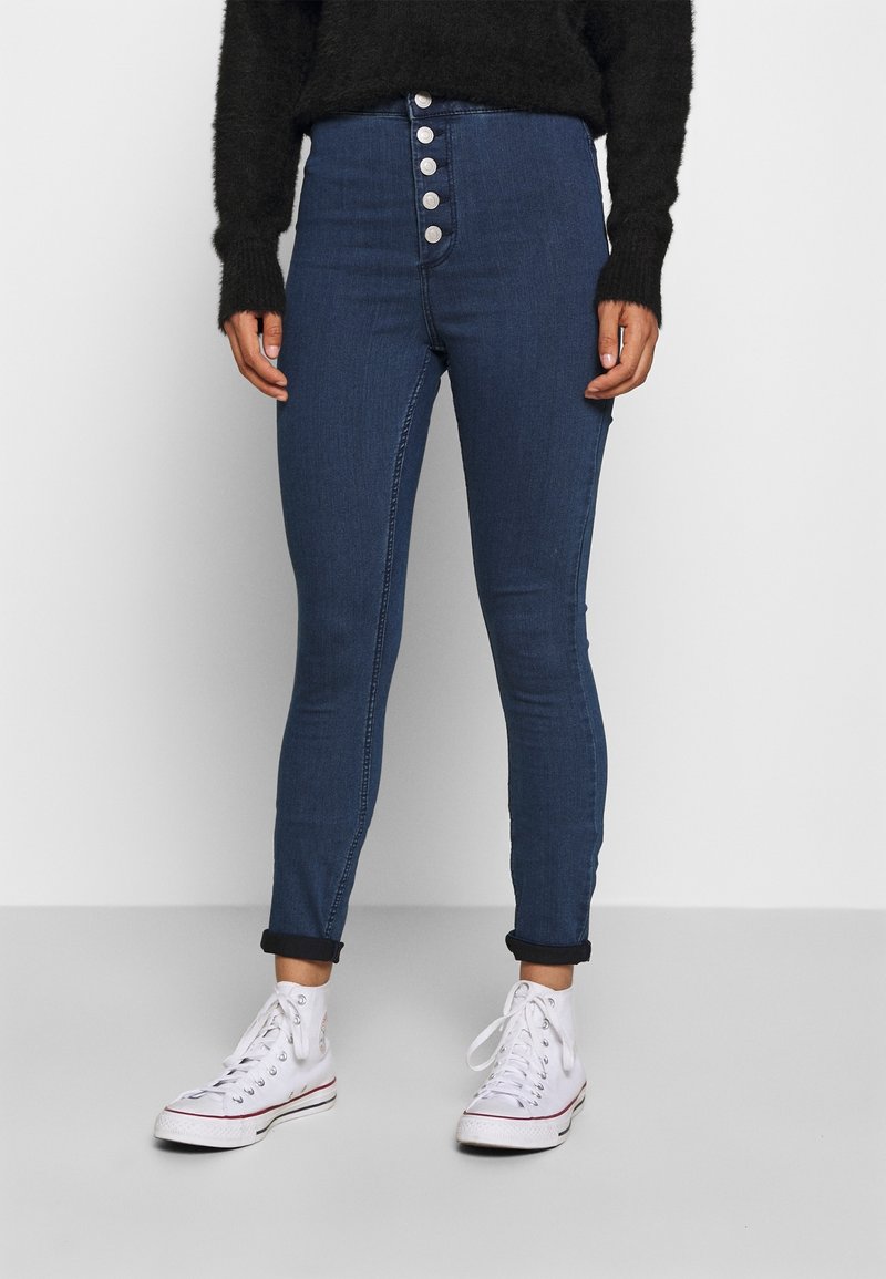 Persona que lleva pantalones ajustados de tiro alto en azul oscuro con cinco botones plateados y zapatillas blancas de caña alta, de pie contra un fondo liso.