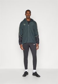 Donkergroene ritsjack met gele accenten, gecombineerd met zwarte slim-fit broek en zwarte sportschoenen, model staat tegen een witte achtergrond.