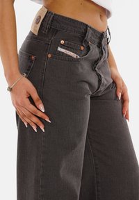 Dark gray denim pants featuring a high waist, decorative stitching, copper rivets, and a small front pocket with a logo patch on the back.