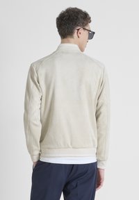 Giacca in suede beige con polsini e vita a costine. Presenta una silhouette bomber classica con una texture liscia e dettagli minimi.
