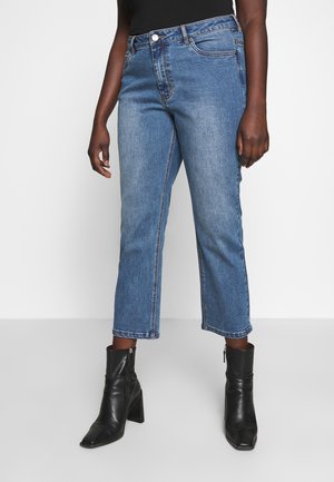 Persoon die mid-rise blauwe cropped jeans en zwarte enkellaarzen met een vierkante neus en hak draagt, staand tegen een effen witte achtergrond.