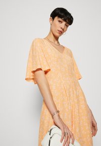 Robe imprimée florale en orange doux avec des manches volantées et un décolleté en V ; assortie avec un sac blanc. Caractéristiques d'un tissu léger et fluide.
