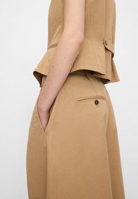 Close-up of a person wearing beige tailored pants and matching vest with hand in side pocket against a plain light background.