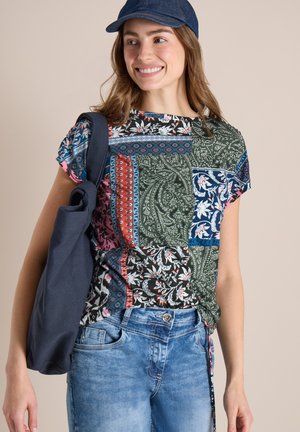 Jeune femme souriante, portant une chemise patchwork à motifs, un jean denim bleu, une casquette marine et un sac fourre-tout marine sur l'épaule.