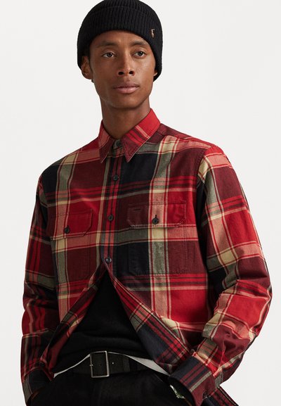 Red and black plaid shirt with button-down collar, two chest pockets, and black buttons. Worn with a black beanie and black belt.
