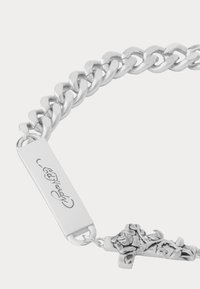 Stříbrný náramek s řetízkem, který má plochou obdélníkovou známku vyrytou s nápisem "Ed Hardy" a křížem zdobeným květinovým designem.