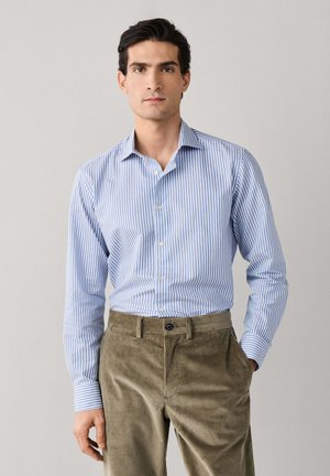 Hombre con camisa de botones a rayas azul claro y blanca metida en pantalones de pana verde oliva, de pie contra un fondo simple.