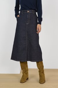 Femme portant une jupe midi en denim foncé avec une couture frontale, un pull bleu marine et des bottes hautes en daim beige, debout sur un sol en bois.