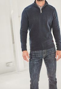 Ein Mann trägt einen navyblauen Quarter-Zip-Pullover über einem karierten Hemd und dunklen blauen Jeans und steht drinnen.