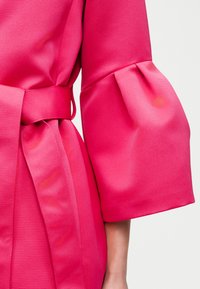 Felroze getextureerde jas met riem en brede, licht opgeblazen mouwmanchet op een arm met een lichte huidskleur.