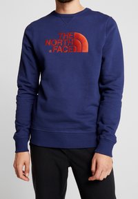 Man som bär en blå sweatshirt med röd "THE NORTH FACE"-logga och svarta byxor, står med avslappnade händer vid sidorna.