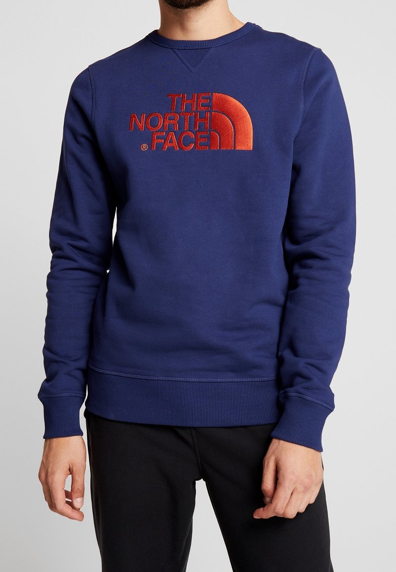 Man som bär en blå sweatshirt med röd "THE NORTH FACE"-logga och svarta byxor, står med avslappnade händer vid sidorna.