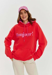 Červená mikina s kapucňou a nápisom "Bonjour!" v ružovej farbe, so šnúrkou na kapucni a prednou vreckom. V kombinácii s ružovou čiapkou a svetlými nohavicami.